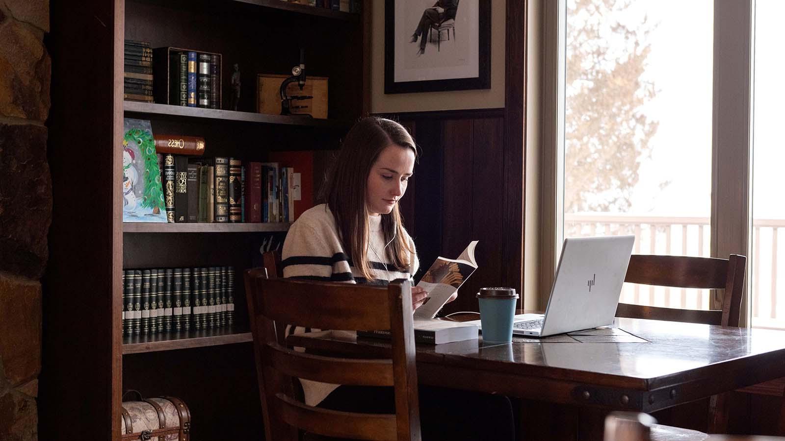 英语 student reading by window with coffee and laptop on table